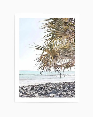 Framed photography art print of a beach scene with a natural wood frame and white border. The composition features a rocky shore in the foreground, with gentle turquoise waves breaking on the sand, and a light blue sky above. A palm tree with long, spiky leaves extends from the right side of the frame, partially obscuring the sky and ocean.