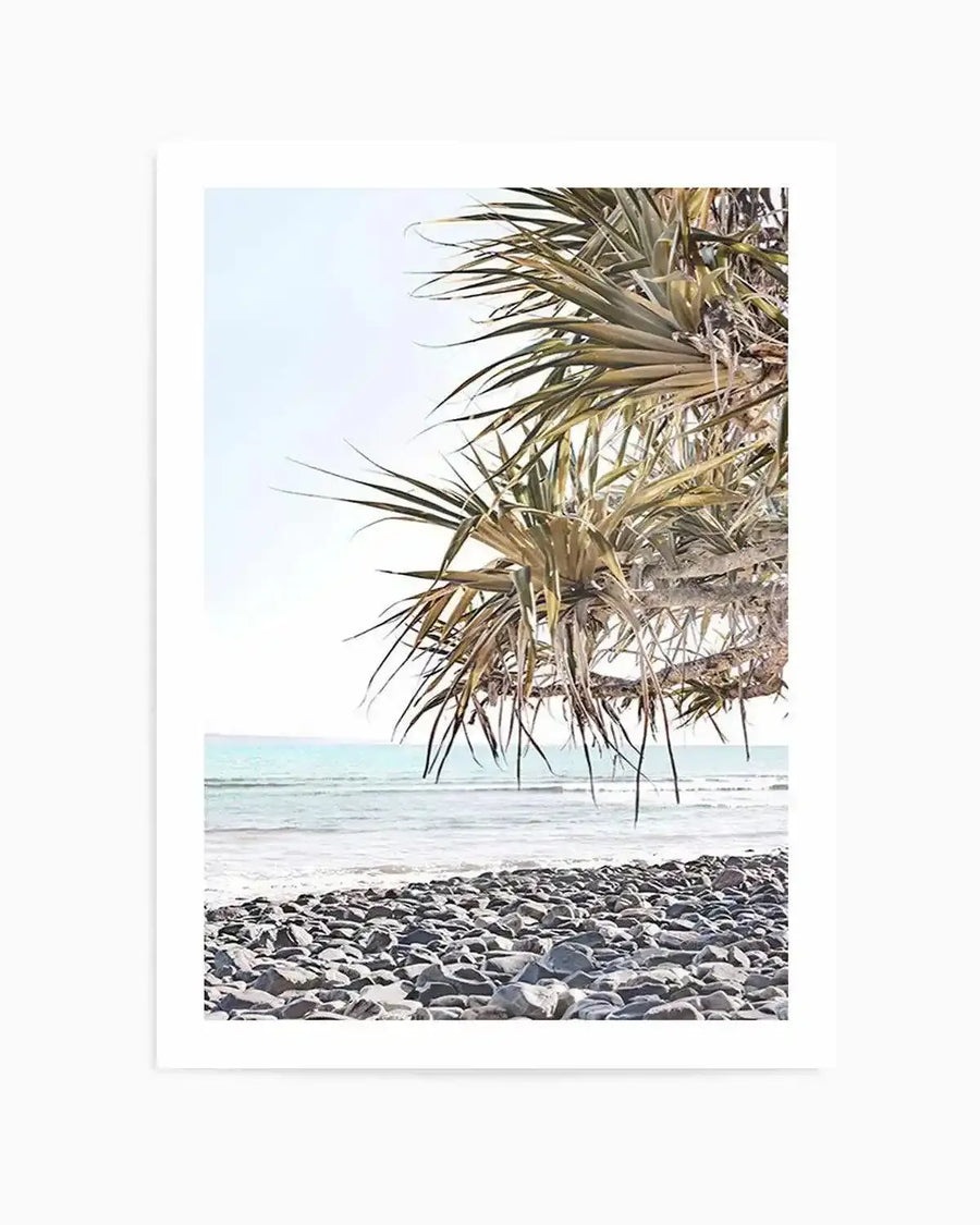 Framed photography art print of a beach scene with a natural wood frame and white border. The composition features a rocky shore in the foreground, with gentle turquoise waves breaking on the sand, and a light blue sky above. A palm tree with long, spiky leaves extends from the right side of the frame, partially obscuring the sky and ocean.