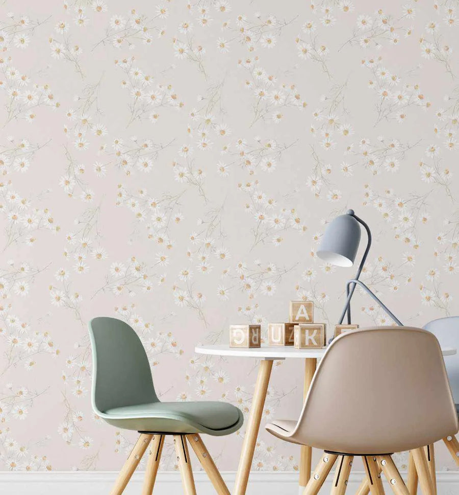 Lifestyle shot featuring Little Daisy Chain wallpaper in a child's play area with a small white table and two chairs. The wallpaper has a repeating pattern of delicate white daisy clusters with subtle yellow centers and thin green stems, set against a soft, pale pink background. Wooden alphabet blocks are scattered on the table, and a light blue desk lamp stands to the right.