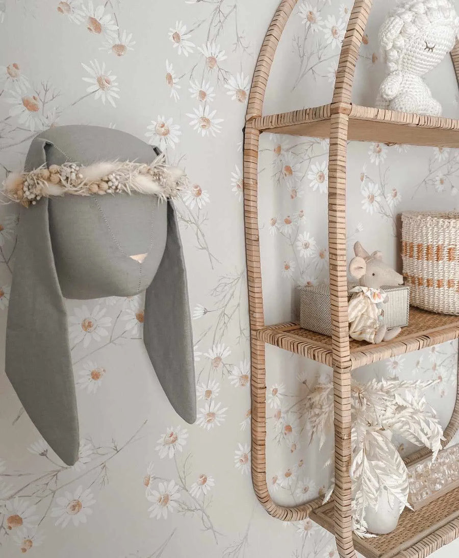 Lifestyle shot of a nursery or child's room featuring Little Daisy Chain wallpaper. The wallpaper has a repeating pattern of hand-painted white daisy flowers with light orange centers and delicate grey-green stems on a soft, muted sage green background. A grey fabric bunny head with long floppy ears and a dried floral crown hangs on the wall, next to a curved rattan shelf displaying a white crocheted lion, a small striped basket, a stuffed mouse, and dried white botanical arrangements in a white vase.