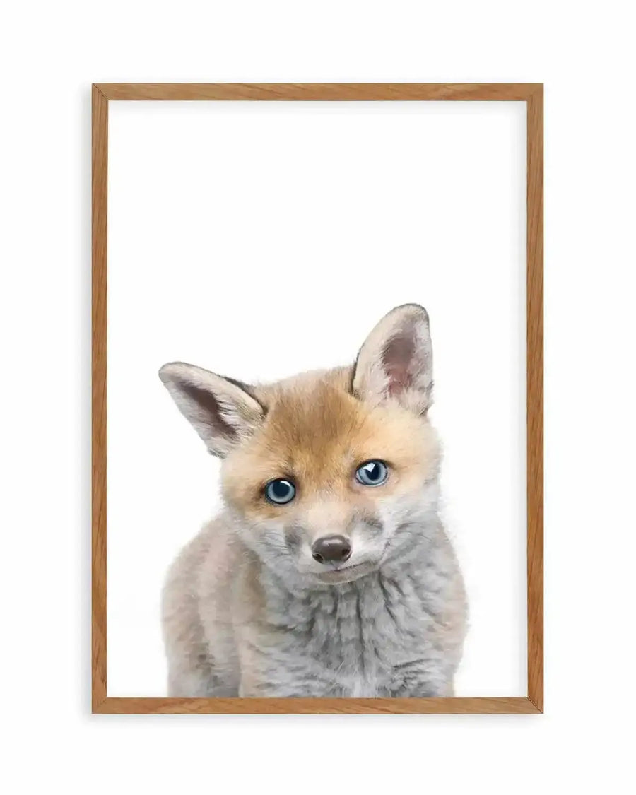 Framed art print featuring a close-up photograph of a baby fox with bright blue eyes and soft, light brown and grey fur, displayed in a natural oak frame with white border. The fox's head is tilted slightly, and it looks directly at the viewer with a curious expression.