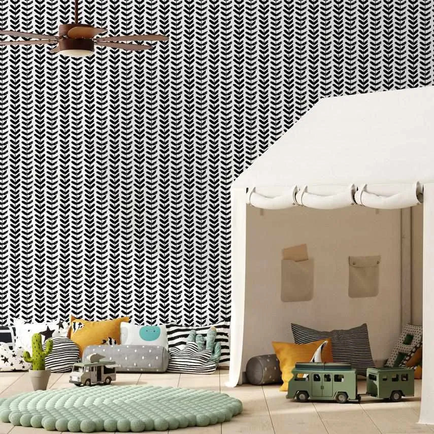 Little Leaf in Black Wallpaper displayed on a child's bedroom wall featuring a geometric herringbone leaf pattern in black and white. The repeating leaf motif creates a vertical striped effect across the wall. The wallpaper is shown in a modern, minimalist room setting with soft sage green and cream accents, a patterned play tent, round textured rug, toy vehicles, and coordinating black and white striped pillows and storage baskets.