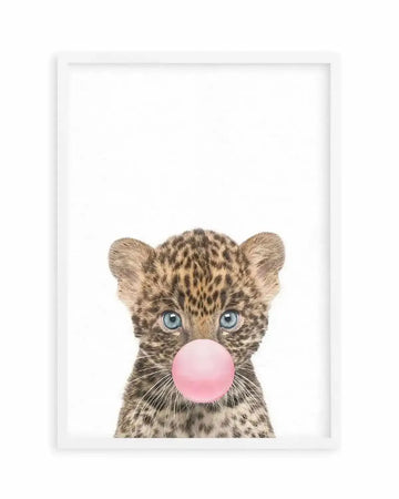 Art print in white frame featuring a young leopard cub with spotted tan and brown fur and bright blue eyes, blowing a large bubble gum bubble in soft pink. The cub's head is centered against a clean white background, rendered in a detailed, photorealistic illustration style. The composition is vertical with ample white space above, creating a minimalist aesthetic. The white frame includes a white border around the artwork.