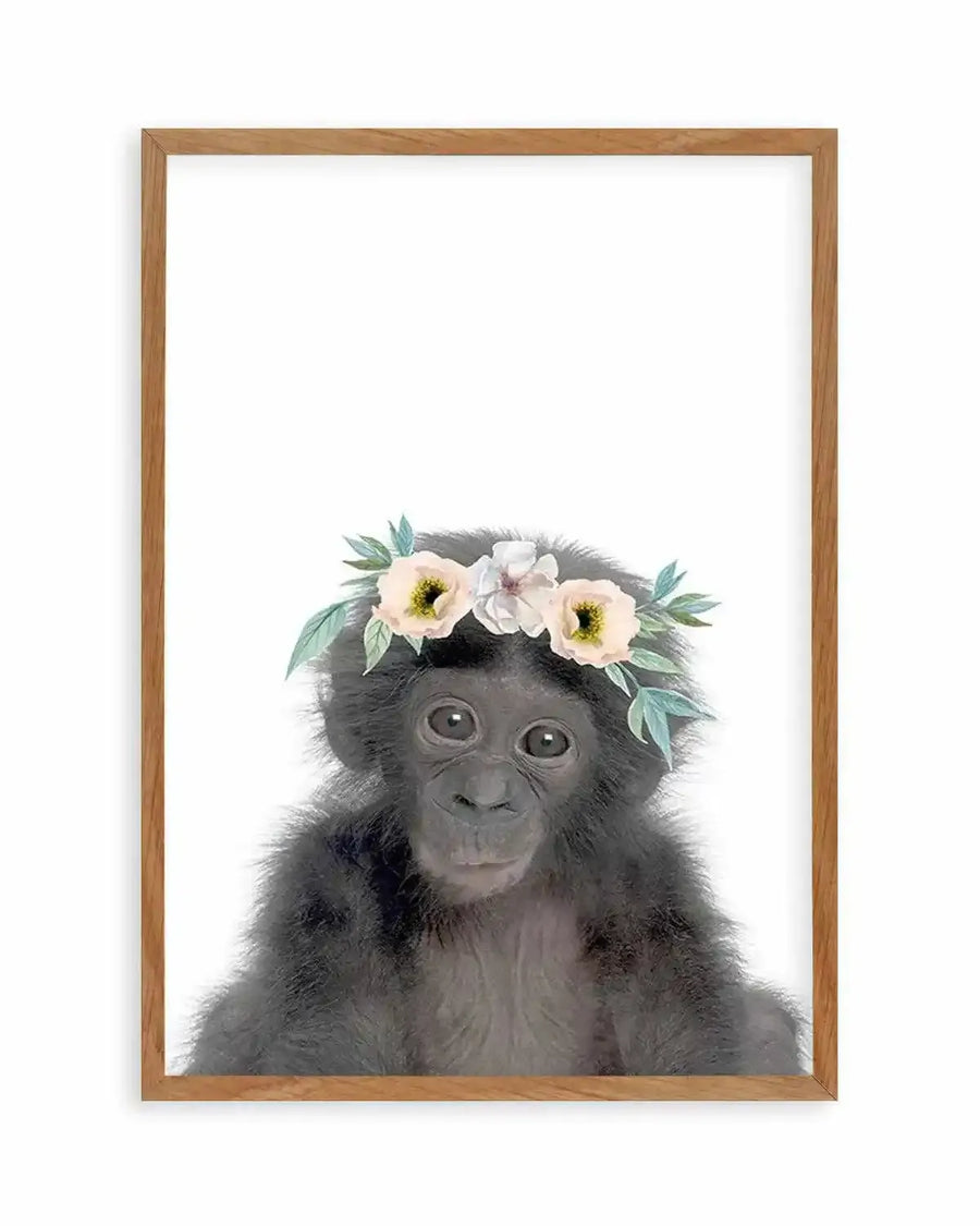 Watercolour art print featuring a baby chimpanzee with dark grey fur and large dark eyes, wearing a flower crown of cream and pale yellow blooms with green leaves, displayed in a natural oak frame with white border.
