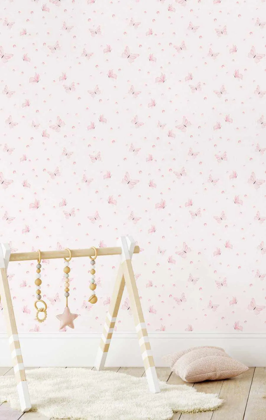Peel and stick wallpaper featuring a repeating pattern of delicate pink butterflies and small pink polka dots on a soft white background. The butterflies have a watercolour quality with subtle shading and varied wing positions, creating a gentle, whimsical design. The wallpaper is shown in a nursery setting with a wooden baby gym and a cream rug.