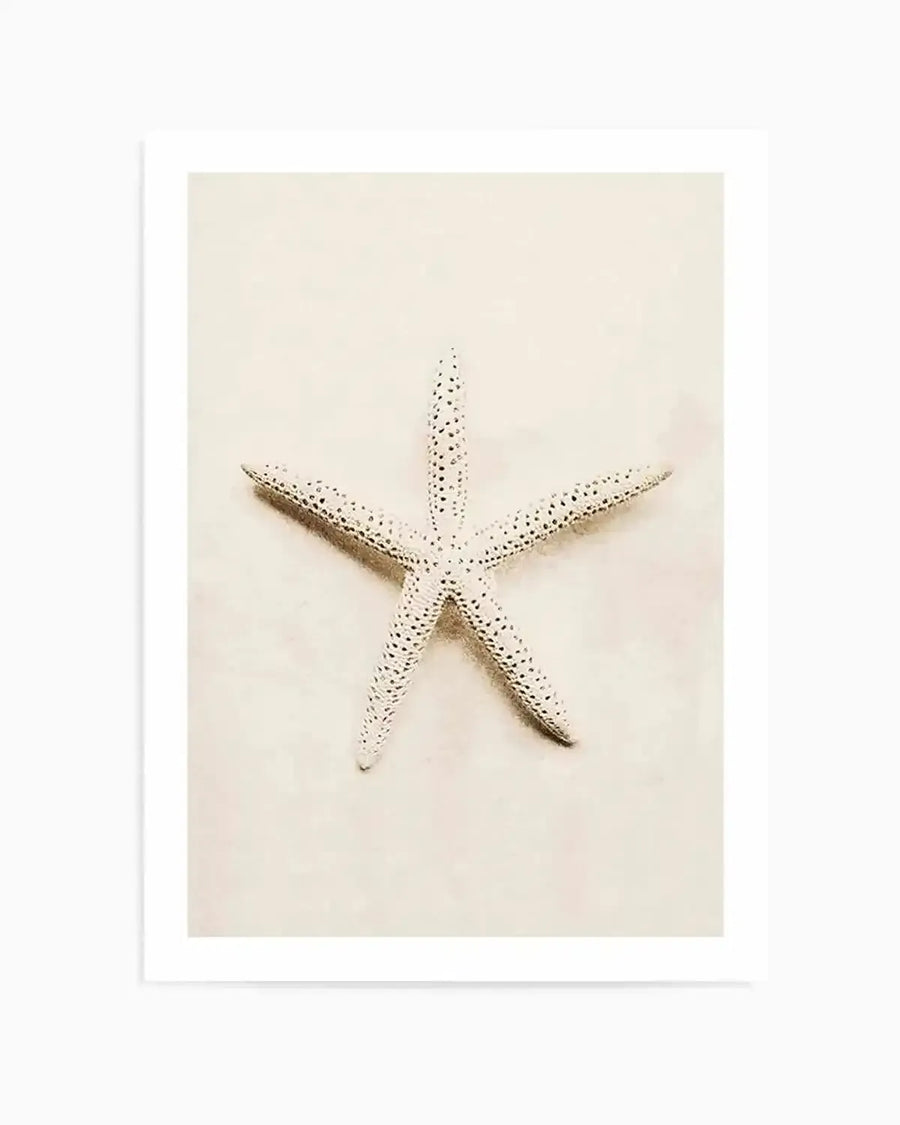 "Framed photography art print featuring a single white starfish with textured bumps, centered on a soft, sandy beige background. The starfish has five arms, each with small, dark dots, and casts a subtle shadow, creating a serene, minimalist composition. The print is displayed with a white border."