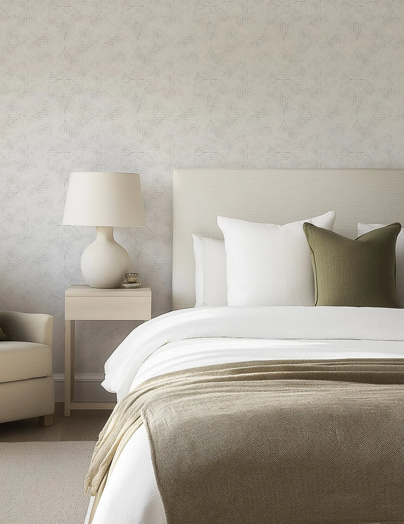"Bedroom featuring a wall covered in Little Arches in Grey Wallpaper, a repeating pattern of interlocking semi-circles in a light grey hue on a soft white background, creating a subtle geometric design. The wallpaper is displayed behind a bed with a cream upholstered headboard, white bedding, and a dark olive green throw pillow. A light wood bedside table with a white ceramic lamp and small decorative bowl sits to the left of the bed, with a cream armchair partially visible."