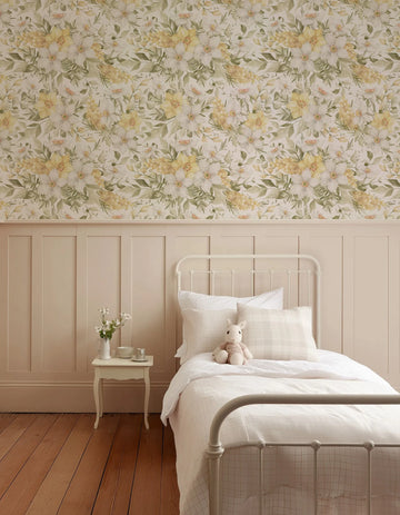 "A child's bedroom featuring Little Golden Fleur Wallpaper on the upper half of the wall, above cream-coloured wainscoting. The wallpaper displays a repeating pattern of hand-painted white and yellow flowers with green leaves and small orange accents on a light cream background. The room includes a white metal bed frame with white bedding, a small white side table with a vase of white flowers, and light brown wooden floorboards."