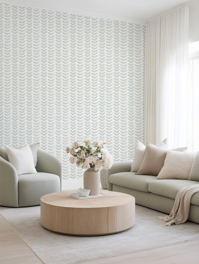 Lifestyle shot of a minimalist living room featuring a wall covered in Little Leaf in Sage wallpaper. The wallpaper has a repeating pattern of hand-painted, muted sage green leaf motifs arranged in vertical rows on a soft white background, creating a subtle botanical texture. The room includes a light sage green sofa with neutral-toned pillows and a throw, a matching armchair, a round light wood coffee table with a vase of white flowers, and sheer white curtains.