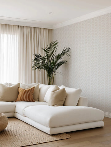 Lifestyle shot of a minimalist living room featuring a white sectional sofa with various throw pillows, a large potted plant, and a wall adorned with "Little Leaf in Sand" wallpaper. The wallpaper features a repeating pattern of small, stylized leaf or chevron shapes in a muted sand or light beige tone on a soft off-white background. The room has light-colored curtains, a woven rug, and light wood flooring, creating a serene and natural aesthetic.