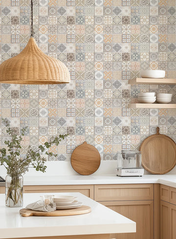 Lifestyle shot of a kitchen featuring Little Moroccan Tiles Wallpaper as a backsplash. The wallpaper has a repeating pattern of small square tiles, each with a unique geometric or floral design in muted tones of beige, brown, grey, blue, and orange. The kitchen has light wood cabinetry, white countertops, and open wooden shelves holding white bowls. A large woven pendant light hangs in the foreground, and a clear vase with green foliage sits on the counter.