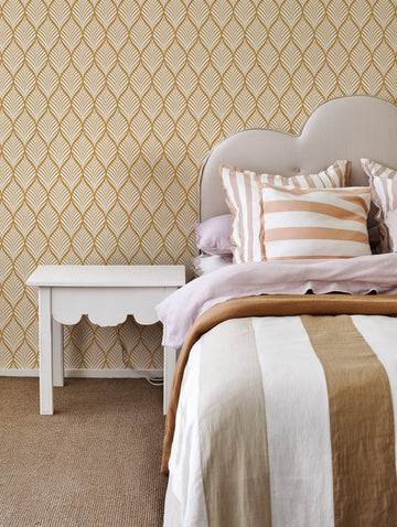 Lifestyle shot of a bedroom featuring Little Retro Leaf Wallpaper in a warm mustard yellow and off-white pattern. The wallpaper covers the wall behind a bed with a curved, light grey upholstered headboard. The bed is dressed with light pink linen bedding, a brown throw, and striped pillows in shades of white, light pink, and brown. A white scalloped bedside table sits to the left of the bed, and the floor is covered with a textured brown carpet.