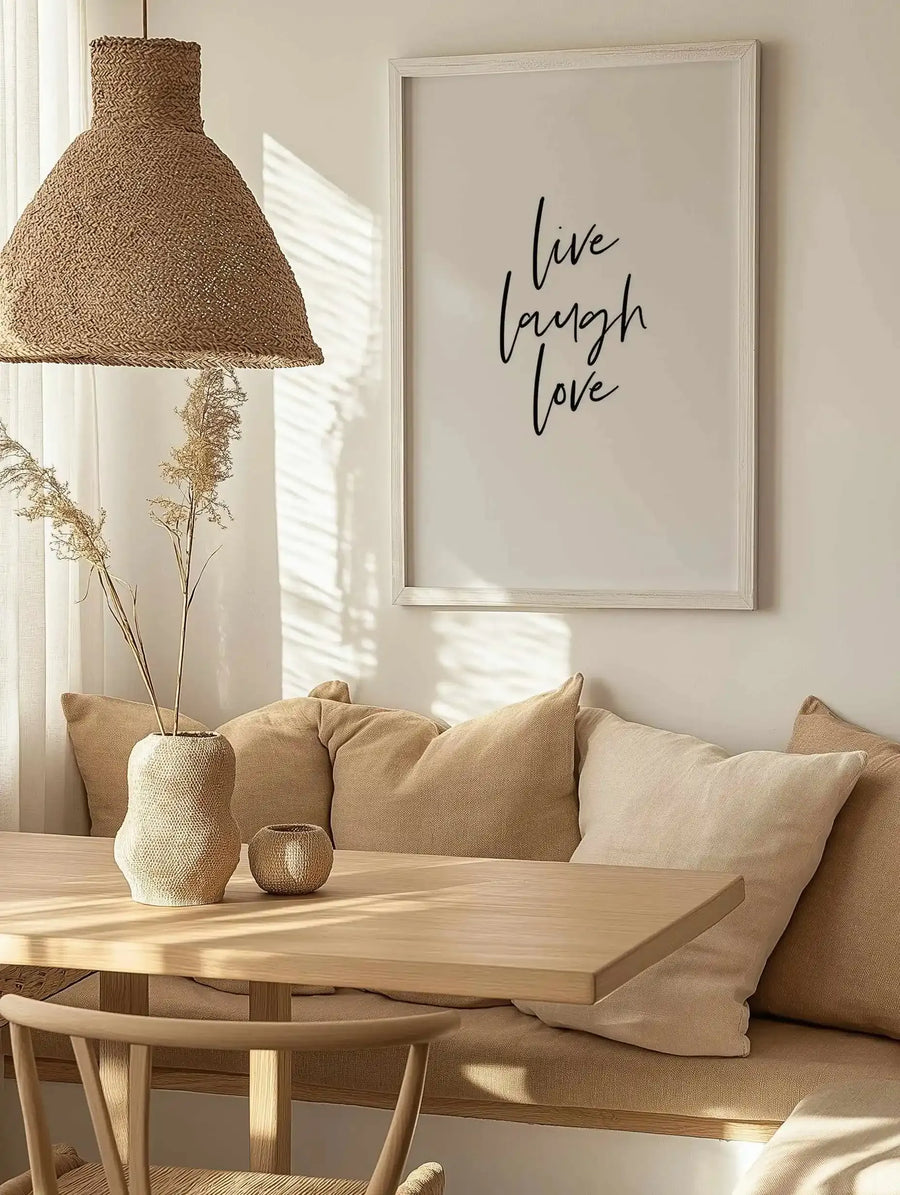 Framed art print displayed on a beige wall above a natural wood table and tan upholstered sofa. The print features handwritten-style black text reading "live laugh love" on a cream background, set in a whitewashed wooden frame. Natural light streams across the scene. A woven pendant lamp, ceramic vase with dried flowers, and neutral linen pillows complete the minimalist, warm aesthetic.