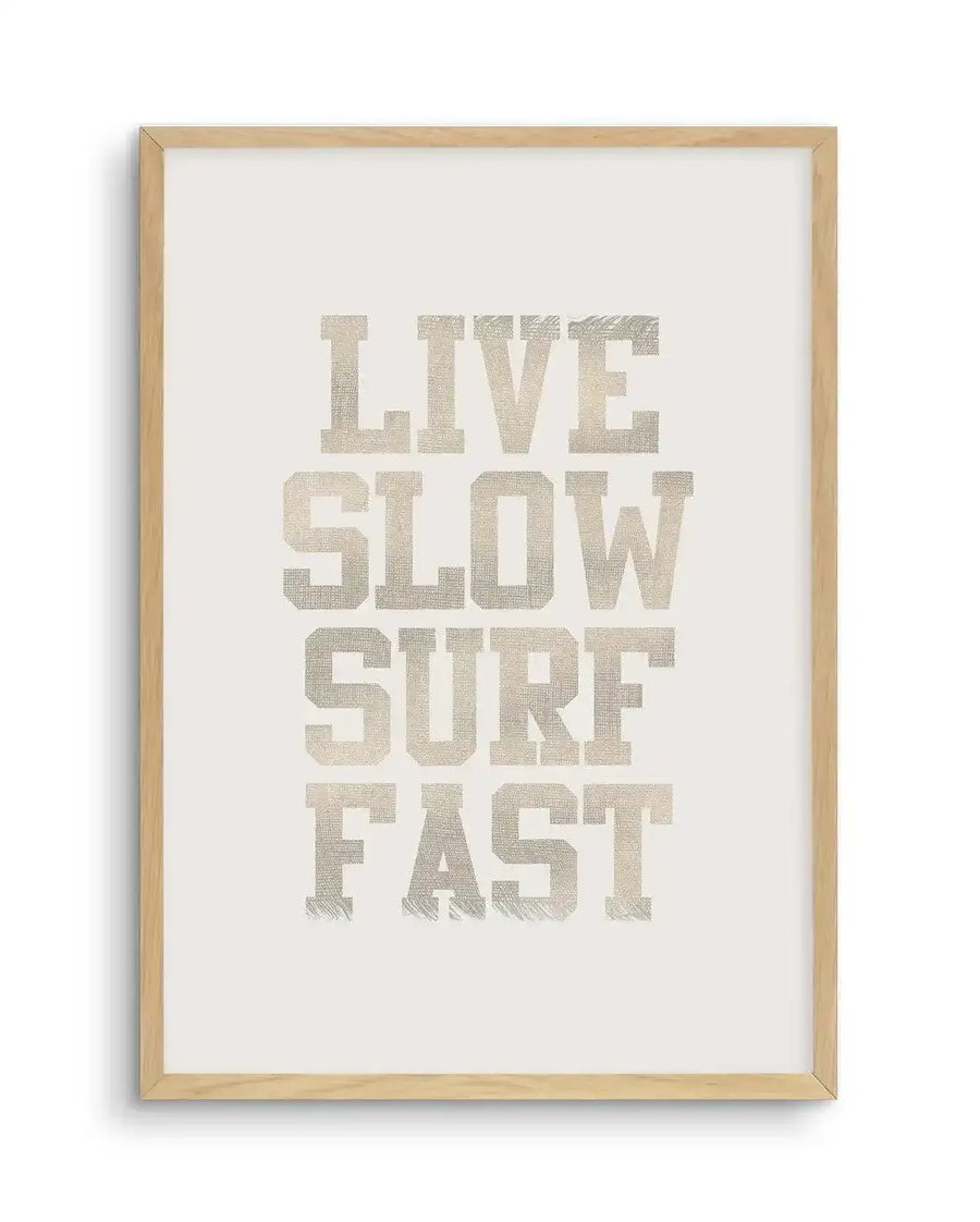 Art print featuring the words "LIVE SLOW SURF FAST" in a bold, uppercase, distressed font with a woven texture, printed in a light beige or off-white colour on a clean white background, displayed in a natural oak frame with white border.