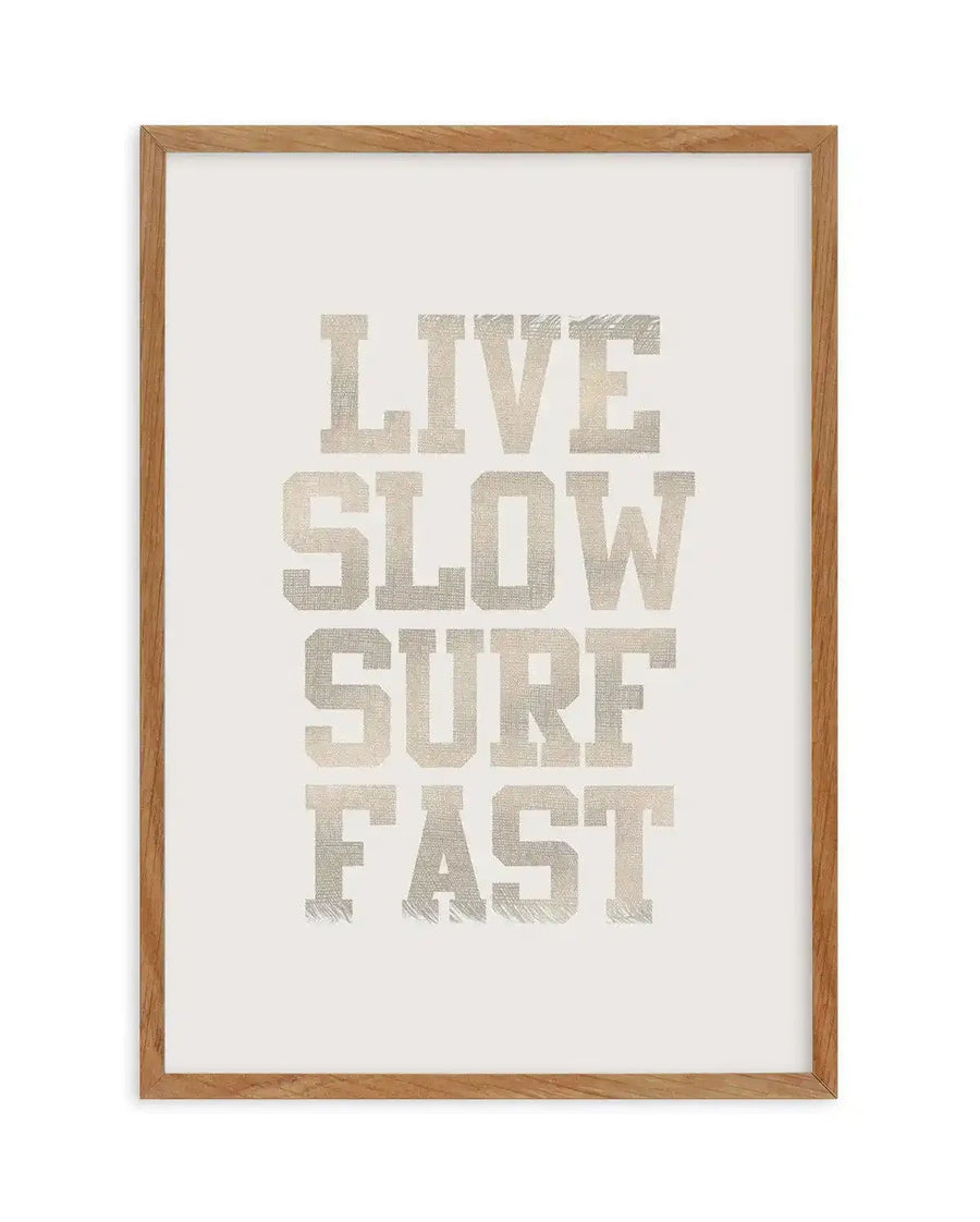 Art print featuring the words "LIVE SLOW SURF FAST" in a distressed, collegiate-style font with a woven texture, printed in a light beige-grey on a white background, displayed in a natural oak frame with white border.