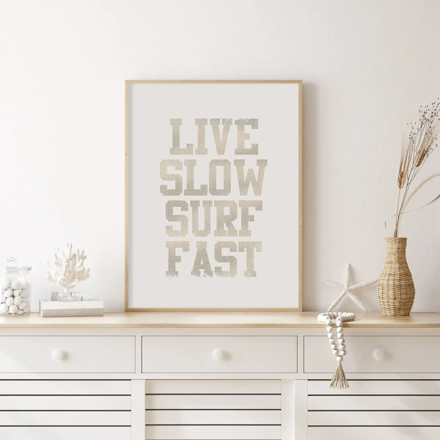 Framed art print displayed above a white wooden dresser in a bright, minimalist living room. The print features the words "LIVE SLOW SURF FAST" in a distressed, textured light beige font on a cream background, with a natural wood frame. The dresser is adorned with coastal-themed decor, including a glass jar of white pebbles, a white coral sculpture, a white starfish, a woven vase with dried wheat, and a string of white wooden beads.