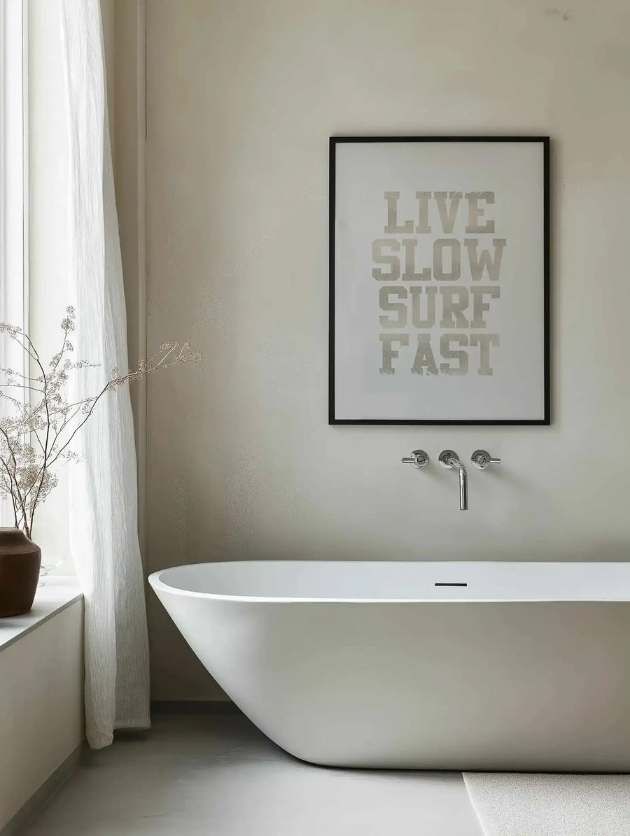 Bathroom:
Framed art print displayed above a freestanding white bathtub in a minimalist bathroom. The print features the words "LIVE SLOW SURF FAST" in distressed grey block letters on a white background, with a black frame and white border. The room includes a sheer white curtain, a brown ceramic vase with dried flowers, and chrome wall-mounted tapware.