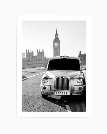 Framed black and white photograph of a classic London taxi cab on a street with the Houses of Parliament and Big Ben in the background, displayed in a white frame with a white border. The taxi is facing forward, slightly angled, with "LONDON" on its license plate and "TAXI" on its roof sign. The sky is bright and clear, and the overall mood is iconic and timeless.