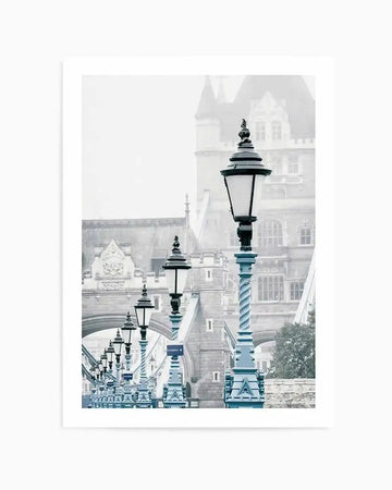 Framed photography art print of London's Tower Bridge on a misty day, with a row of blue lampposts in the foreground. The composition features the iconic bridge in soft grey tones, partially obscured by fog, with the ornate blue lampposts creating a leading line towards the distant towers. The mood is serene and atmospheric.