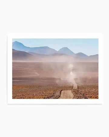"Framed photograph of a desert landscape with a dirt road and mountains in the distance, with a natural wood frame and white border. The composition features a vehicle creating a large dust cloud as it drives along a winding dirt road through a vast, arid plain with sparse, low-lying vegetation. In the background, layers of blue-grey mountains rise under a pale blue sky."