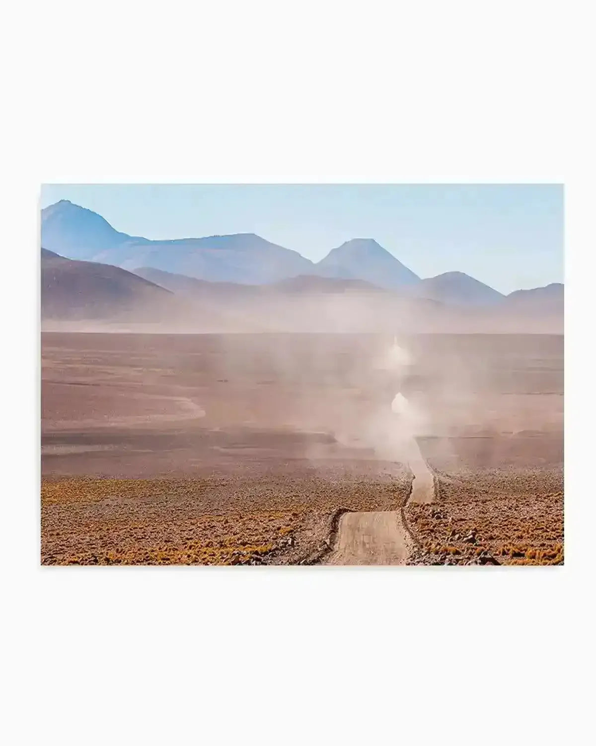 "Photography art print of a long, winding dirt road in a vast, arid landscape under a clear blue sky. The road stretches into the distance, with a plume of dust rising from it, suggesting a vehicle has recently passed or is approaching. Mountains with soft blue and purple hues line the horizon, and the foreground is covered in sparse, golden-brown vegetation."