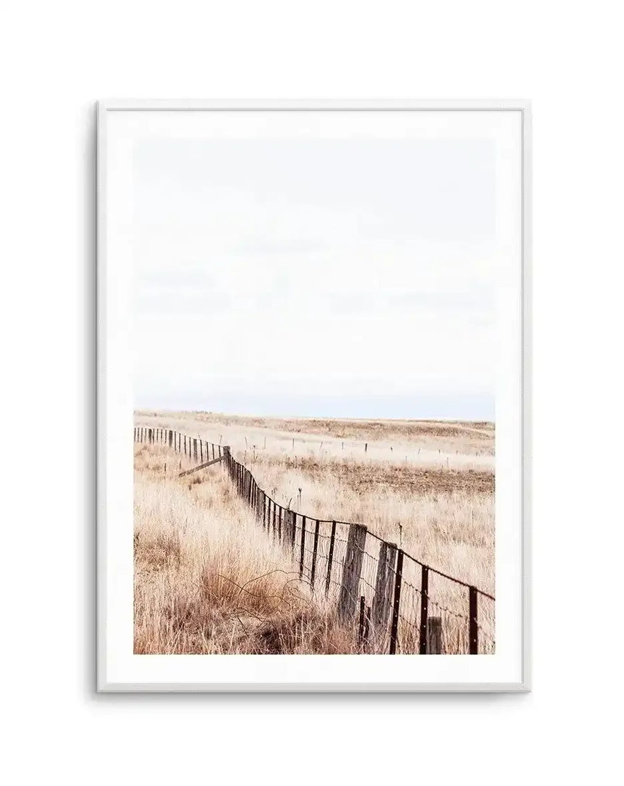 Framed art print titled "Long Road Home" showing a landscape photograph with a weathered wooden fence running diagonally through dry, golden-brown grassland. The fence recedes into the distance across rolling terrain with sparse vegetation and a calm blue-grey sky above. The photograph has warm, muted earth tones of tan, rust, and ochre. Presented in a light grey frame with white border.
