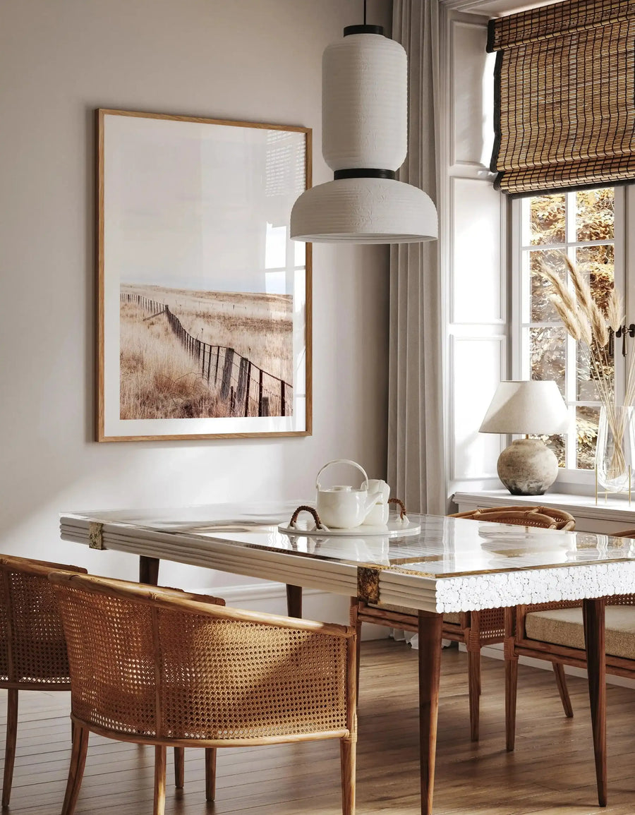 Long Road Home art print displayed in a natural wood frame with white border, hung on a beige wall above a marble-topped desk. The photograph shows a sandy, overgrown field with dried grasses and a wooden fence line receding into a hazy, muted landscape in warm beige and brown tones. The print is styled in a bright, minimalist workspace with woven rattan chairs, cream pendant lights, and a woven roman shade, creating a calm, natural aesthetic.