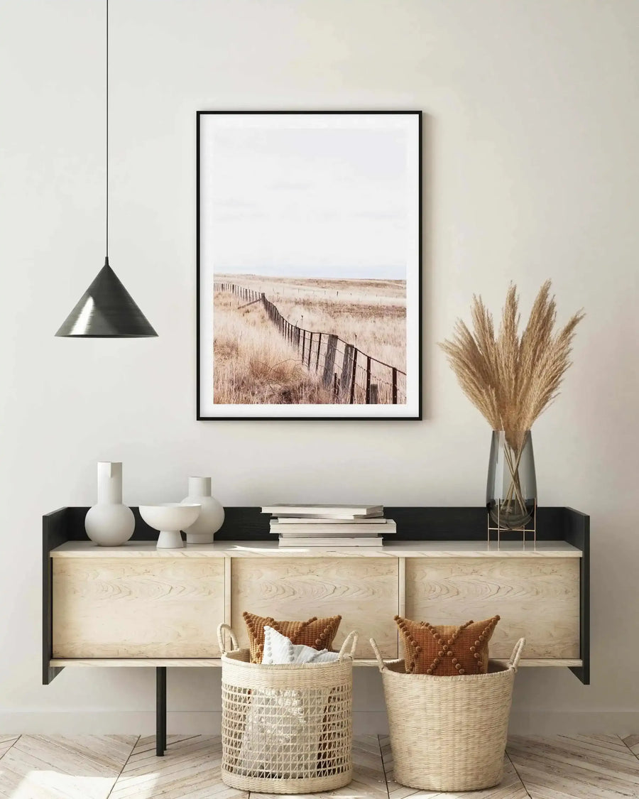 Lifestyle shot of a minimalist living room featuring a framed art print of a long fence in a dry, grassy field. The print is a photograph with a muted, natural color palette of light browns, creams, and pale blues under a bright, overcast sky, displayed in a black frame with a white border. The print hangs above a light wood and black console table adorned with white ceramic vases, a stack of books, and a tall glass vase with dried pampas grass. Two woven baskets with throw pillows sit on the floor below.