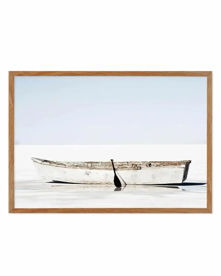 Framed photograph of a weathered white rowboat with a single oar, resting on a calm, reflective body of water under a pale blue sky, displayed in a natural oak frame with white border.