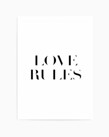 Art print featuring the words "LOVE RULES" in large, bold black serif capital letters with geometric cutout details on a white background. The typography has a modern, minimalist aesthetic with clean lines and negative space within certain letterforms. The print is displayed on a white or off-white background with a subtle gray border visible on the left edge, suggesting a framed presentation.