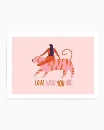 Art print featuring a stylized illustration of a woman with long dark hair riding a pink tiger with orange stripes, on a pale pink background. The text "LOVE WHO YOU ARE" is written in orange at the bottom.