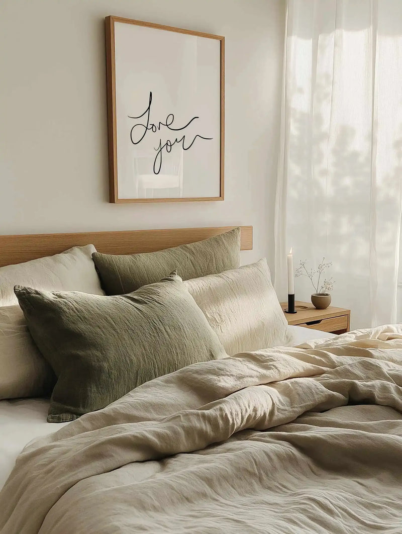 Framed art print displayed above a wooden bed headboard in a minimalist bedroom. The print features "Love you" written in flowing, hand-scripted black cursive lettering on a cream background, set in a natural wood frame with white border. The bedroom includes sage green and cream textured pillows, beige linen bedding, a wooden nightstand with a small ceramic vase and dark candle, and sheer white curtains filtering soft natural light.
