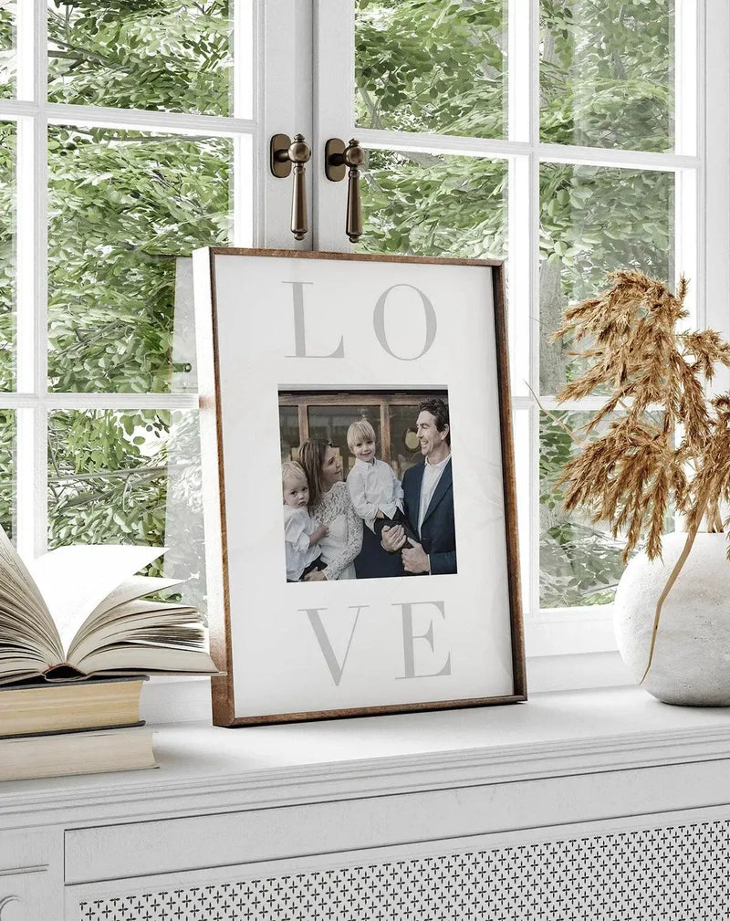 Framed art print displayed on a white windowsill featuring the word "LOVE" in large, light gray serif letters arranged vertically, with a black and white family photo of two adults and two children centered between the L and VE. The print has a white background with a rustic wood frame in warm brown tones. The windowsill styling includes an open book, stacked books, a white textured vase with dried pampas grass, and large multi-pane windows with green foliage visible outside.