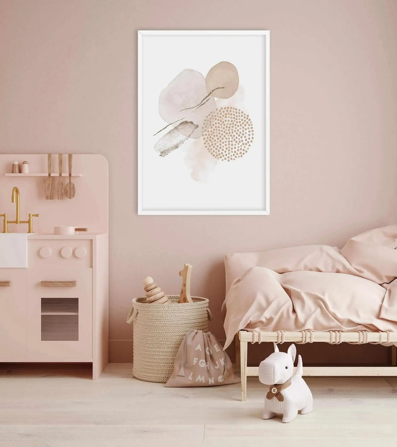 Nursery:
Framed abstract art print displayed above a bed in a child's bedroom. The print features organic shapes in soft blush pink, beige, and gold with speckled and brushed textures, set against a white background with a white frame. The room includes a light pink play kitchen, a woven basket with wooden toys, a beige storage bag with white letters, and a white striped dog plush toy.