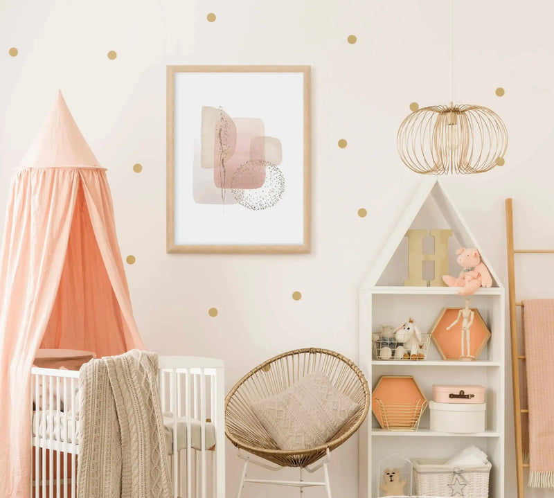 Nursery:
Framed art print displayed above a white crib with a peach canopy in a baby's nursery. The print features abstract watercolour shapes in dusty rose, blush pink, and cream, with delicate gold glitter accents, printed on fine art paper and displayed in a natural oak frame with white border. The room includes a woven chair with a cream pillow, a white bookshelf with various decorative items, and a gold wire pendant light.