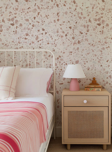 Terrazzo wallpaper featuring irregular, scattered fragments in shades of dusty rose, terracotta, and pale grey on a light cream background, displayed on a bedroom wall behind a white metal bed frame. The bed is dressed with white sheets, a white pillow with red striped trim, and a striped pink and white throw blanket. A light wood nightstand with a cane-front drawer and a pink lamp sits beside the bed.