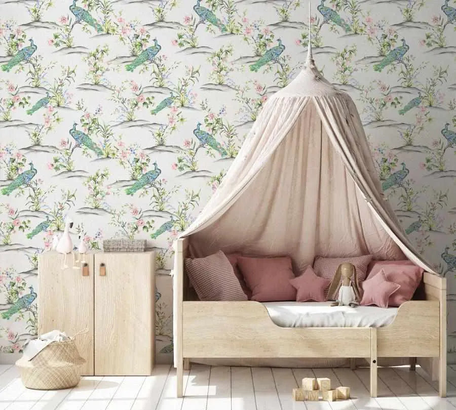 Nursery:
"A child's bedroom featuring a light wood bed with a draped canopy in soft beige, positioned against a wallpapered wall. The wallpaper features a repeating pattern of peacocks with blue and green plumage perched on branches with delicate pink and purple flowers and green leaves, set against a light cream background. A light wood cabinet with two doors and a woven basket are visible on the left, and wooden blocks are on the floor in front of the bed."