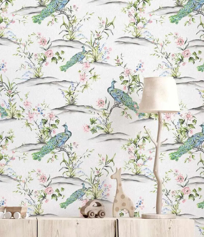 Luxe Peacock Wallpaper displayed on a bedroom wall featuring a repeating pattern of iridescent peacocks with blue-green plumage perched among rolling grey hills and scattered pink roses, blue flowers, and green foliage in watercolour illustration style on a cream background. A white floor lamp and wooden toy car sit on a light wood shelf in front of the wallpaper.
