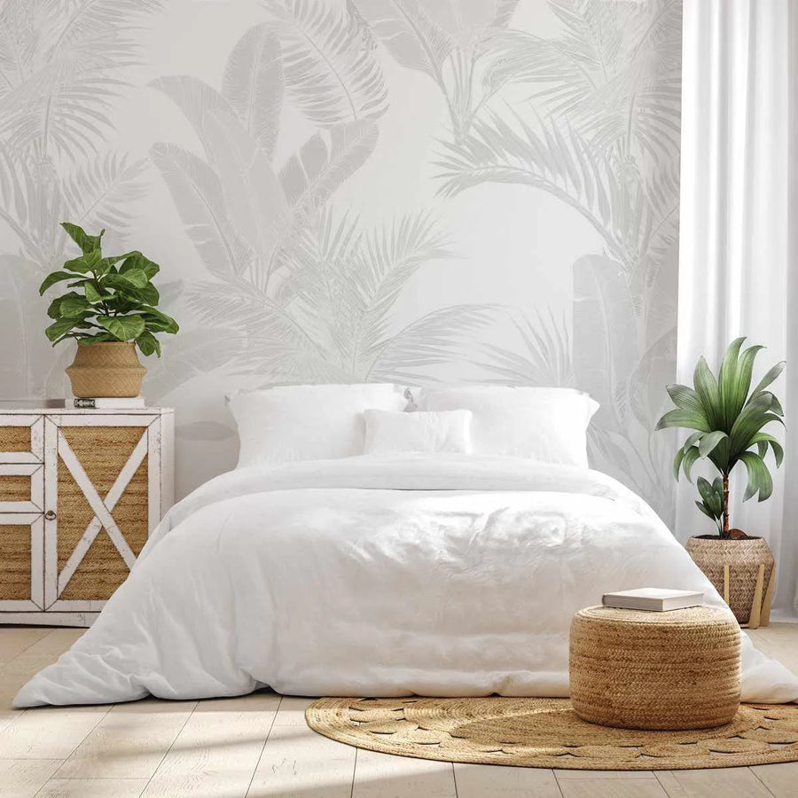 Luxe Tropical in Grey Wallpaper displayed on a bedroom wall behind a white bed. The wallpaper features large-scale palm and fern fronds in soft grey and beige tones on a cream background, creating a botanical pattern with subtle depth. The room is styled with natural woven furniture, potted green plants, a rattan pouf, and light wood flooring, demonstrating the wallpaper's tropical aesthetic in a serene, minimalist bedroom setting.