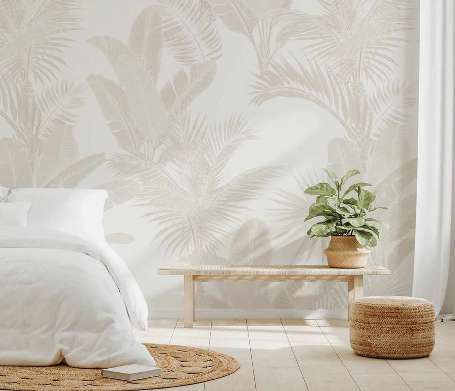 Lifestyle shot of a bedroom featuring Luxe Tropical in Sand wallpaper. The wallpaper covers a full wall and displays a repeating pattern of large-scale tropical leaves, including palm fronds and banana leaves, in a muted sand or beige tone against a soft white background. The room includes a bed with white bedding on the left, a light wood bench with a woven top, a potted plant in a woven basket, and a round woven jute rug on a light wood floor. A white curtain is visible on the right.