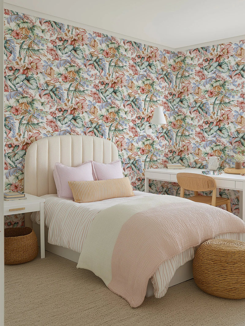 "A bedroom with a bed and desk, featuring a wall covered in Luxe Floral Palm Wallpaper. The wallpaper has a repeating pattern of large-scale tropical flowers and palm leaves in a watercolour style, with colours including dusty rose, peach, sage green, light blue, and warm brown on a soft white background. The bed has a cream upholstered headboard, pink and striped yellow pillows, and a striped white and pink duvet. A white desk with a woven chair and a white nightstand with a woven basket are also visible."