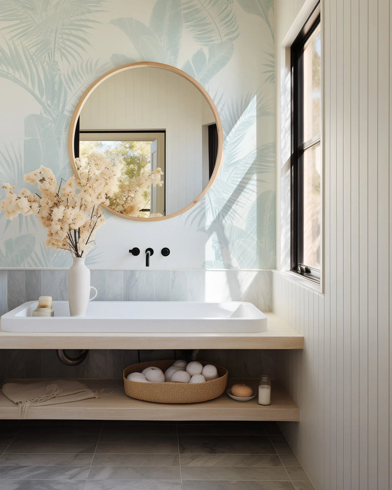 Bathroom:
Bathroom with light blue tropical leaf wallpaper, a round wooden mirror, and a white sink. The wallpaper features large, stylized palm and banana leaves in a soft azure blue against a creamy white background. A white vase with dried fluffy branches sits on the counter next to the sink. The room has a light wood vanity, white vertical paneling, and a window with a black frame.