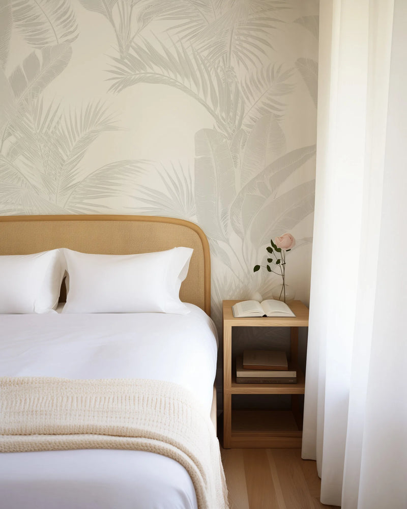 "Luxe Tropical in Grey wallpaper featuring a repeating pattern of large-scale tropical leaves in a light grey outline on a warm cream background, displayed behind a bed in a bright bedroom. The room includes a bed with a woven cane headboard, white bedding, a cream knitted throw, and a wooden bedside table with an open book and a single pink rose in a clear vase."