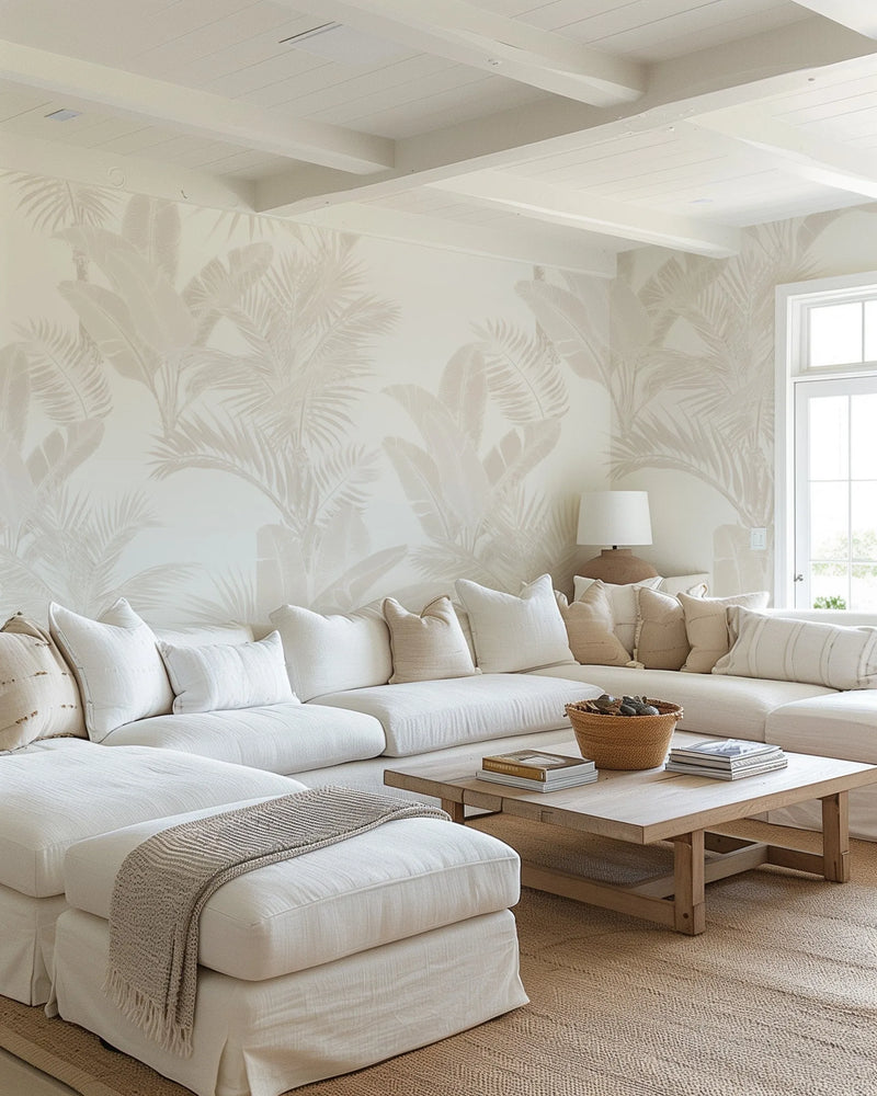 Lifestyle shot of a living room featuring a large white sectional sofa and a light wood coffee table on a natural fiber rug. The walls are adorned with peel and stick wallpaper featuring a repeating pattern of large-scale tropical palm and banana leaves in a muted sand or beige tone on a soft cream background. The room is bright and airy with a large window and door on the right, and the ceiling has exposed white beams.