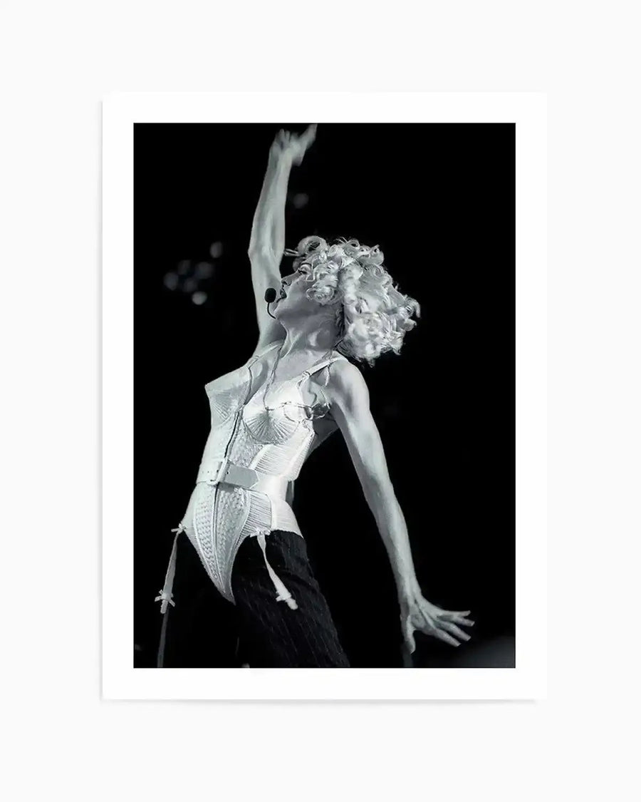 Framed black and white photograph of Madonna performing on stage, captured mid-movement with her right arm raised high. She wears a structured white corset and dark trousers, with curly blonde hair and a headset microphone. The background is dark with soft, out-of-focus lights, creating a dramatic and energetic mood.