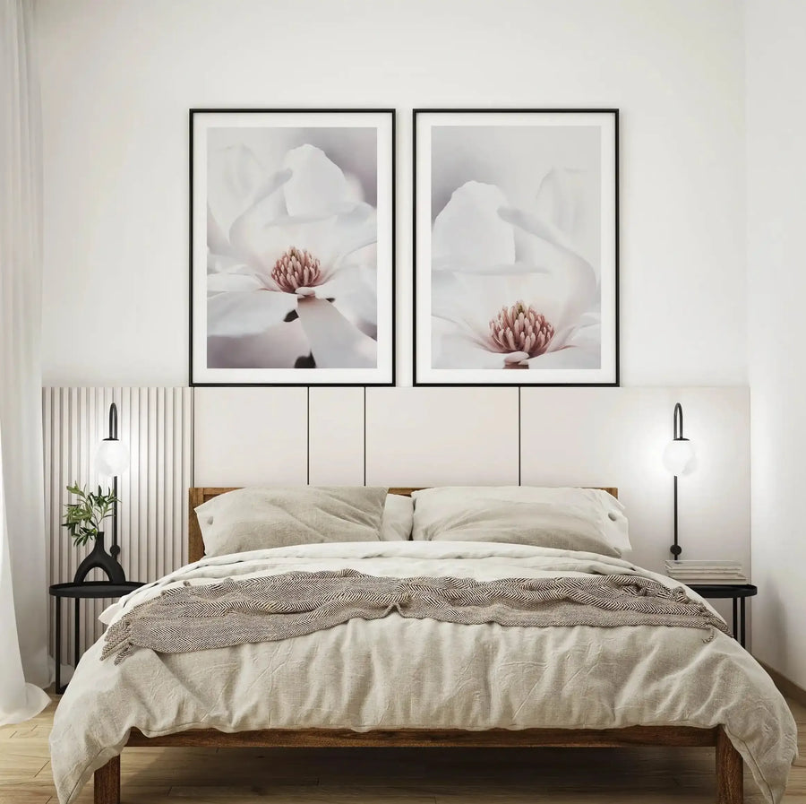 Lifestyle shot of a minimalist bedroom featuring two framed art prints of a white magnolia flower with a pink center, displayed above a bed with a light wood frame and cream bedding. The prints are framed in black with a white border. The room has white walls, a fluted white panel on the left, and two black wall sconces with spherical white glass shades.