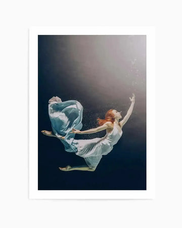 "Art print featuring an underwater photograph of a woman with long red hair, wearing a white slip dress and a flowing light blue fabric, gracefully ascending towards the surface. The deep blue water transitions to a lighter, hazy grey at the top where small bubbles rise. The woman's arms are outstretched, reaching upwards, and her body is angled as if floating towards the light."