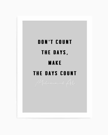 Art print with light grey background featuring black sans-serif text reading "DON'T COUNT THE DAYS, MAKE THE DAYS COUNT" centered on the page. Below the text, a signature in white script reads "Muhammad Ali." The print is mounted in a white frame with a white border.