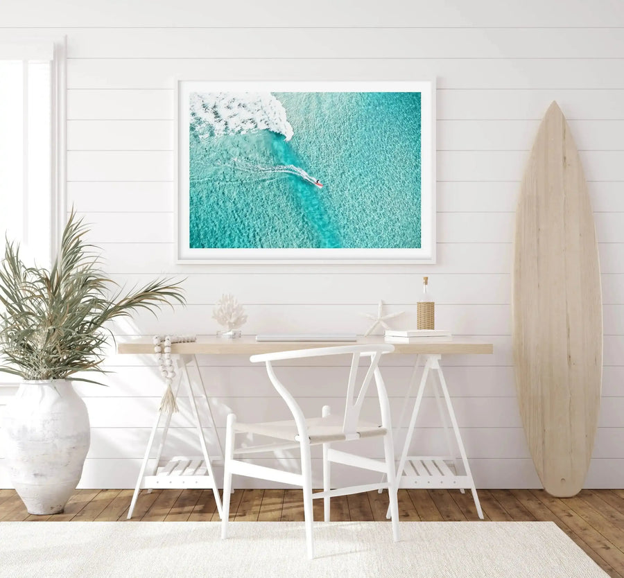 Aerial photograph of a surfer riding a wave at Bondi Beach, featuring turquoise ocean water, white foam, and a small figure on a surfboard. The image is printed on paper and displayed in a white frame with white border, hung on a white shiplap wall above a light wood desk with white chairs. A natural wood surfboard leans against the wall to the right, and a white textured vase with green foliage sits on the left side of the desk.