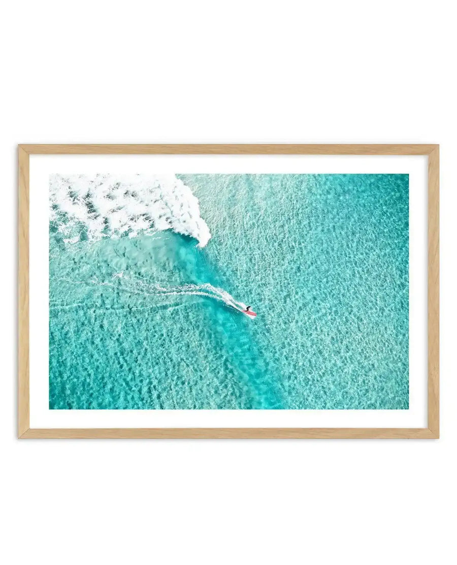 Framed aerial photograph of a surfer on a red surfboard riding a wave at Bondi Beach. The image captures turquoise ocean water with white wave foam and the surfer creating a wake trail. Natural light wood frame with white border. The photograph uses vibrant teals, whites, and soft aqua tones, shot from directly above to show the contrast between the textured water surface and the surfer's movement through the wave.